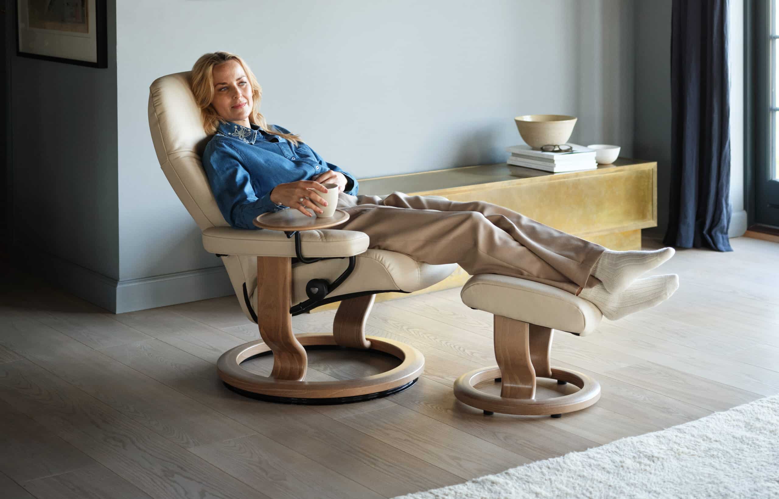 Eine Frau sitzt entspannt in einem Stressless Consul M Classic Ledersessel in der Farbe Creme mit Eichenholzrahmen. Sie trägt einen blauen Jeanshemd, bequeme beige Hosen und weiße Socken. Der Sessel verfügt über eine passende Fußstütze und ein kleines, an den Armlehnen befestigtes Tischchen, auf dem sie eine Tasse hält. Im Hintergrund befindet sich ein niedriger, goldfarbener Schrank mit darauf liegenden Büchern und Dekorationsgegenständen. Der Raum ist modern eingerichtet und strahlt eine gemütliche Atmosphäre aus.