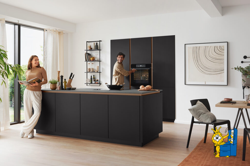 Modern eingerichtete, offene Küche mit dunkler Kücheninsel und Hochschränken in Matt-Schwarz. Ein Mann bedient den Backofen und lächelt, während eine Frau entspannt an der Kücheninsel steht und ein Kochbuch hält. Helle, lichtdurchflutete Atmosphäre mit dekorativen Elementen wie Pflanzen, Wandbild und offener Regalwand. Im Vordergrund rechts ist das Möbel Homann-Maskottchen, der Löwe mit der blauen Kommode, sichtbar.