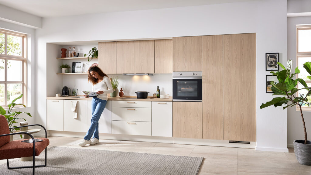 Helle, freundliche Einbauküche mit matten weißen Fronten und Oberschränken in Holzoptik. Eine Frau steht entspannt mit einem Kochbuch in der Hand vor der Küchenzeile. Die Küche ist modern und minimalistisch eingerichtet mit offenen Regalen, Pflanzen und Deko-Elementen. Große Fenster sorgen für viel Tageslicht, im Vordergrund ein gemütlicher Sessel und ein Teppich.