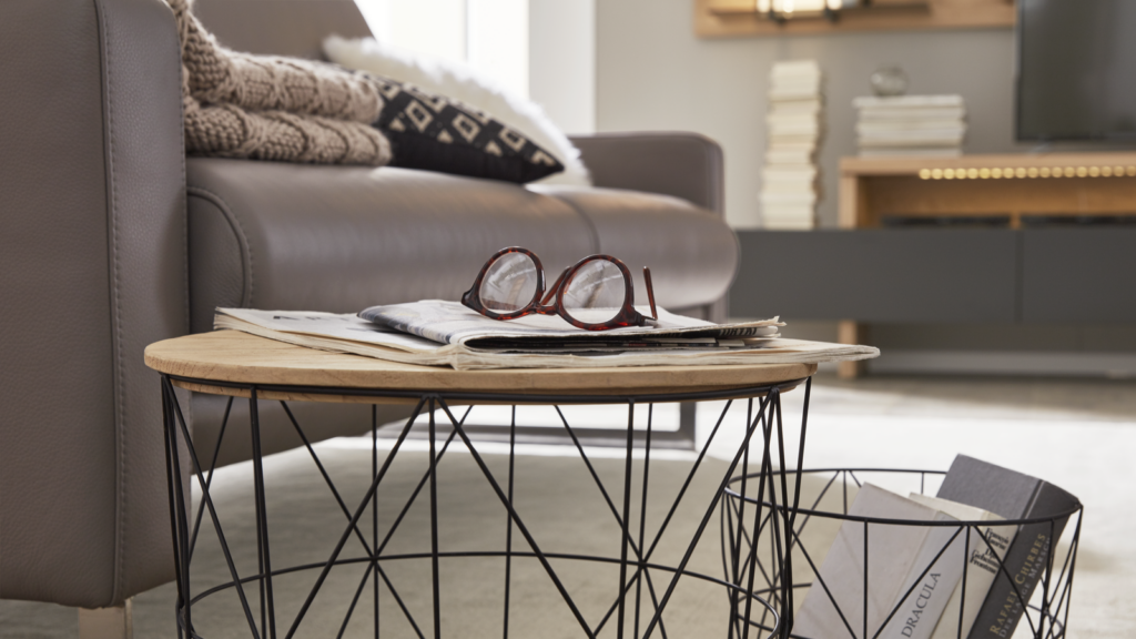 Modern gestaltetes Wohnzimmer mit einem runden Couchtisch mit hölzerner Oberfläche und schwarzem Drahtgestell, auf dem eine Brille, eine Zeitung und eine Zeitschrift liegen. Rechts daneben steht ein Drahtkorb mit mehreren Büchern und Magazinen. Im Hintergrund befindet sich ein graues Ledersofa mit Zierkissen und einer gestrickten Decke, hinter dem ein Regal mit gestapelten Büchern und ein Fernseher zu sehen sind. Der Raum ist durch große Fenster hell ausgeleuchtet und wirkt ordentlich, einladend und entspannt.