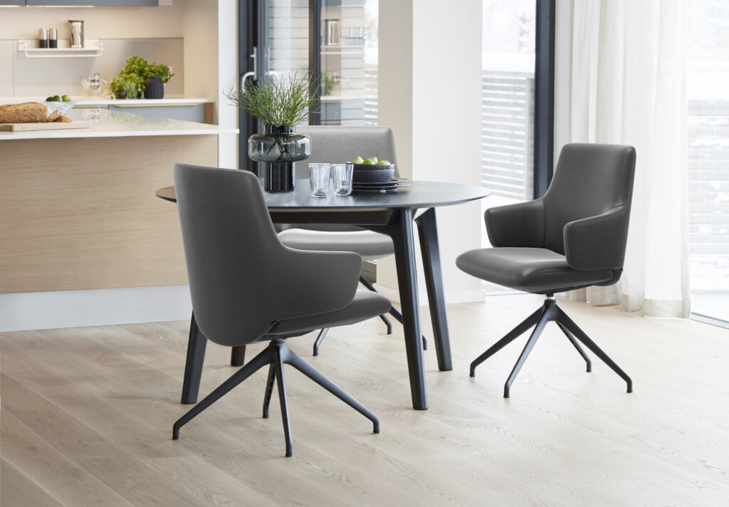 Ein stilvoll eingerichtetes Wohnzimmer mit einem beigen Sofa, dekoriert mit dezenten Kissen. Vor dem Sofa steht ein runder Couchtisch mit schwarzem Metallgestell und einer gläsernen Oberfläche, darauf einige Bücher und eine Kerze. An der Wand hängen moderne, gerahmte Kunstwerke, und im Hintergrund ist eine schlichte Holzkonsole mit Dekoration zu sehen. Der Raum wird von natürlichem Licht durchflutet und wirkt hell und einladend.