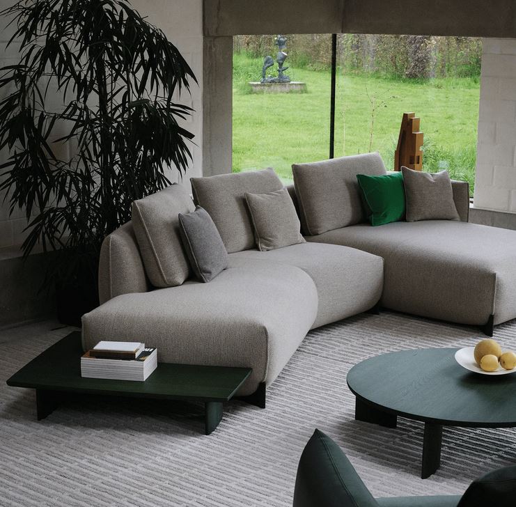 Modernes Wohnzimmer mit einer großen, hellgrauen Eckcouch, geschmückt mit Kissen in verschiedenen Grautönen und einem grünen Kissen. Vor der Couch stehen zwei niedrige, dunkelgrüne Couchtische – einer mit einem Bücherstapel, der andere mit einem weißen Teller und drei gelben Früchten. Ein hoher, schlanker Zimmerpflanze steht links von der Couch. Durch ein großes Fenster ist ein grüner Rasen mit einer Skulptur zu sehen. Das Design ist minimalistisch und modern, mit neutralen Farben und klaren Linien