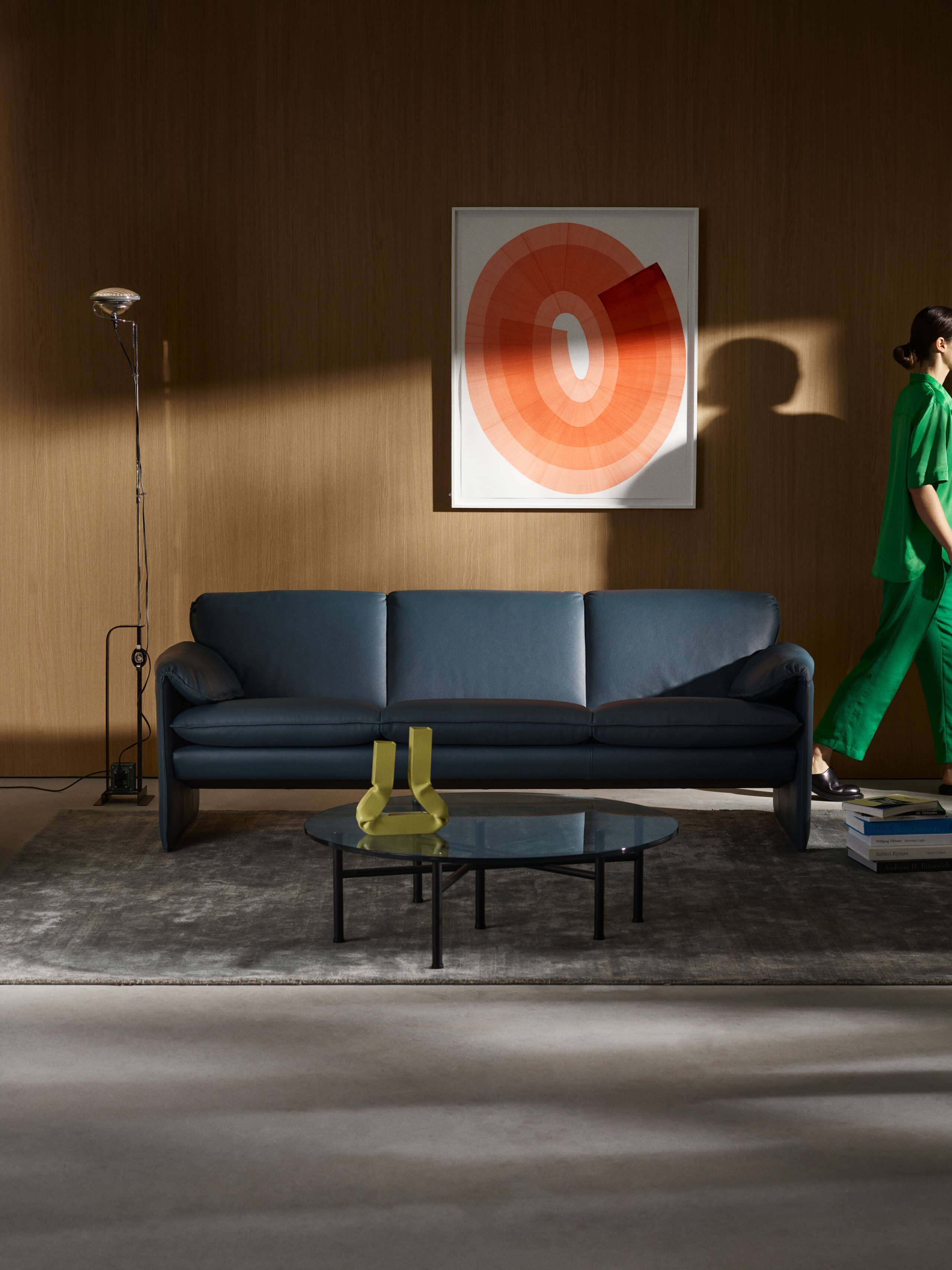 Ein modern eingerichtetes Wohnzimmer mit einer dunklen blauen Couch vor einer Holzvertäfelung. Über der Couch hängt ein gerahmtes Kunstwerk mit konzentrischen Kreisen in warmen Orange- und Rottönen. Links steht eine schlanke Stehlampe. Vor der Couch befindet sich ein runder Glastisch mit einem gelben Dekorationsobjekt. Rechts im Bild geht eine Person in grüner Kleidung aus dem Raum. Der Boden ist mit einem grauen Teppich bedeckt, und neben der Person liegen einige gestapelte Bücher