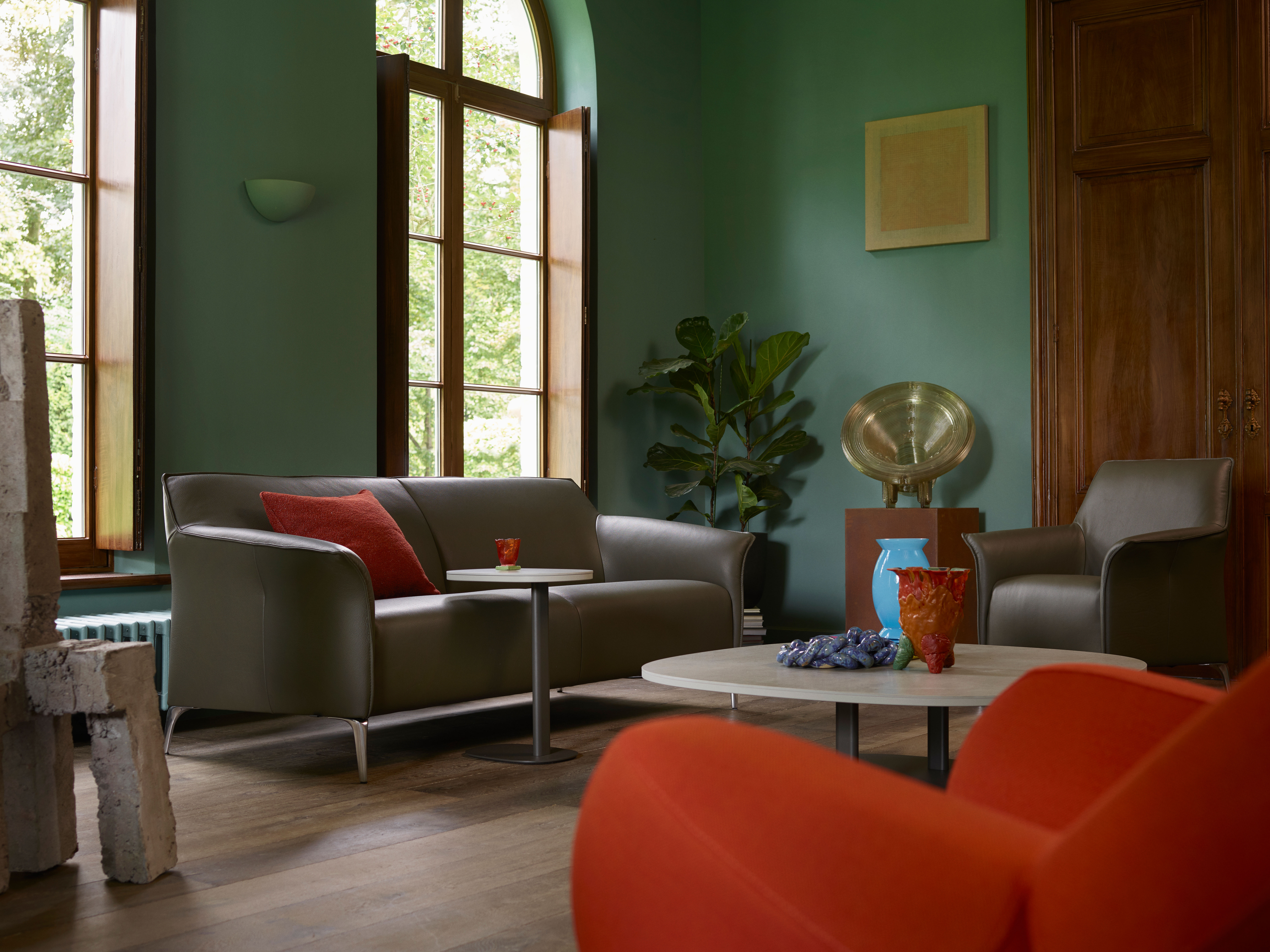 Eine gemütlich eingerichtete Wohnstube mit grünen Wänden und großen Fenstern, die Tageslicht hereinlassen. Die Einrichtung umfasst ein graues Sofa mit einem roten Kissen, einen kleinen runden Beistelltisch mit einem roten Glas darauf und einen grauen Sessel. In der Mitte steht ein runder Couchtisch mit dekorativen Elementen, darunter eine blaue Vase und eine rote Schale. Eine große Pflanze steht in der Ecke, während ein messingfarbenes Dekorationsstück auf einem Holzständer präsentiert wird. Der Raum besitzt einen Holzboden und rechts ist eine Holztür sichtbar. Die Atmosphäre wirkt insgesamt einladend und stilvoll mit einer Mischung aus modernen und klassischen Elementen