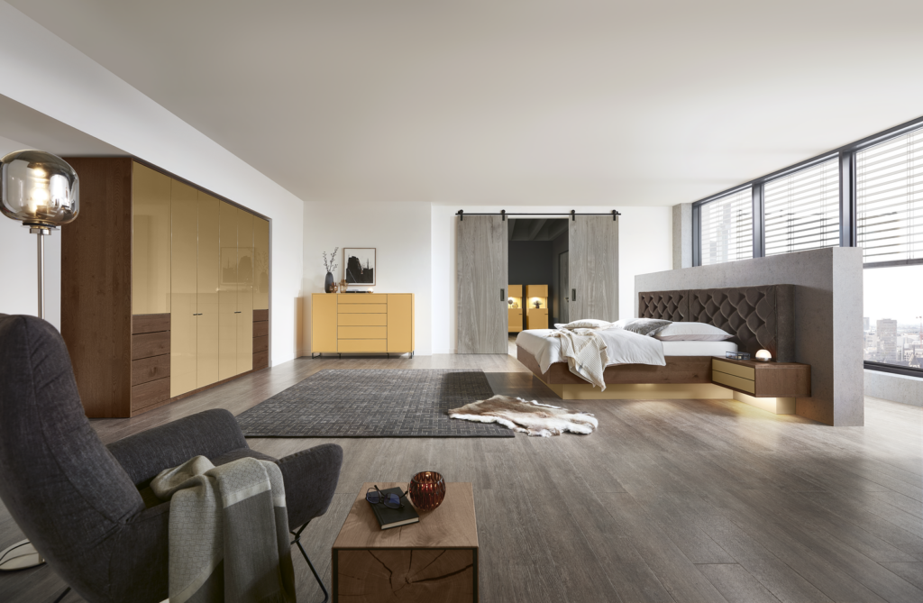Modernes Schlafzimmer mit großem Bett und grauem Polsterkopfteil, flankiert von zwei Holz-Nachttischen. Im Vordergrund ein grauer Sessel mit Überwurfdecke neben einem kleinen Beistelltisch. Ein Teppich in Grau bedeckt den Holzboden. Im Hintergrund ein Kleiderschrank mit gelb abgesetzten Holztüren, daneben eine Holzkommode mit Dekoobjekten. Große Fenster mit Lamellen lassen Tageslicht herein, links führen Schiebetüren im Scheunenstil in einen weiteren Raum