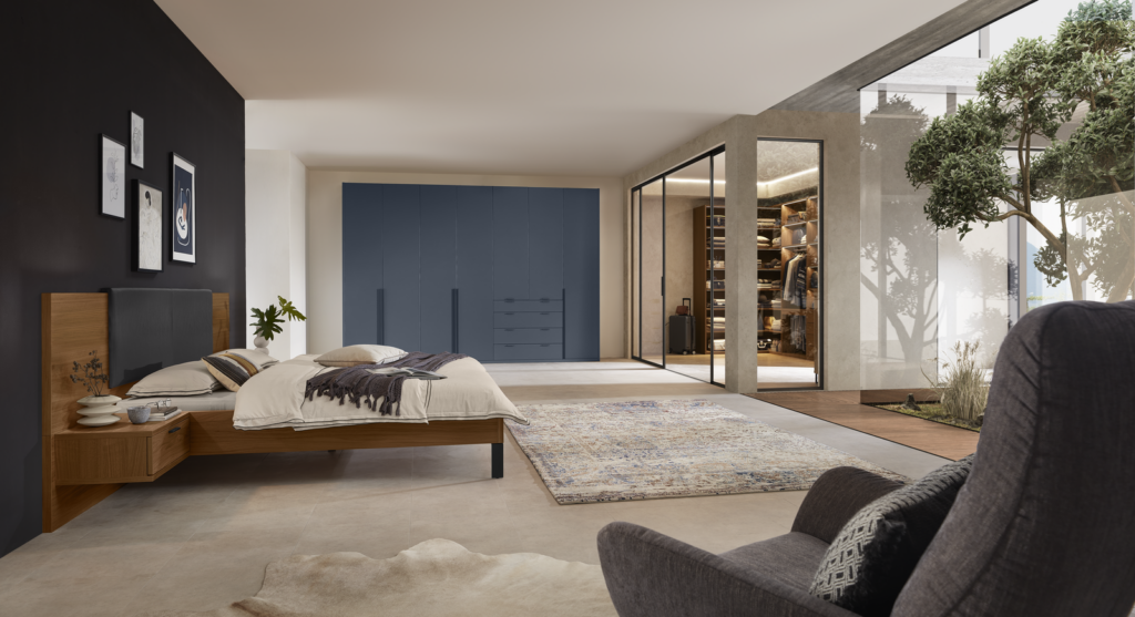 Modernes Schlafzimmer mit großer Holz-Bettanlage und dunklem Kopfteil vor einer schwarzen Akzentwand mit gerahmten Bildern. Rechts ein offener begehbarer Kleiderschrank mit Glasfronten, ordentlich sortierter Kleidung und Regalen. Tageslicht fällt durch hohe Fenster ein, vor denen ein Baum zu sehen ist. Im Vordergrund ein gemütlicher Sessel auf einem gemusterten Teppich – klare Linien und wohnliche Eleganz