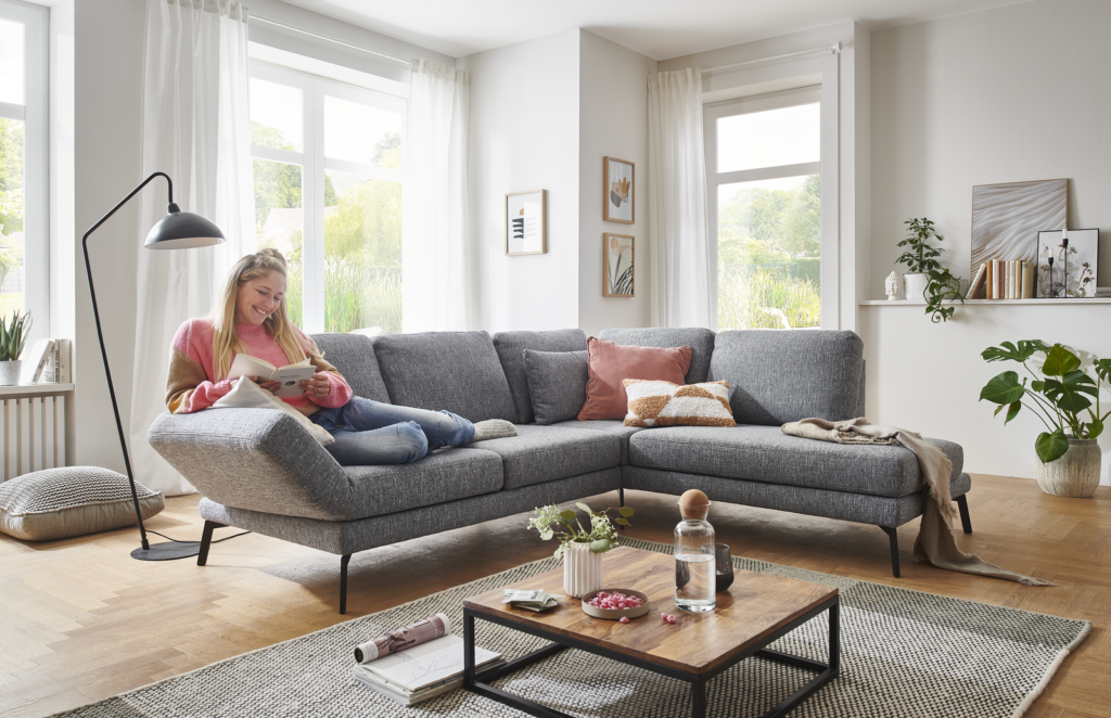 Modern eingerichtetes Wohnzimmer mit großem grauen Ecksofa, auf dem eine Person sitzt und ein Buch liest. Der Raum ist von natürlichem Licht durch große Fenster mit weißen Vorhängen durchflutet. Neben dem Sofa steht eine schwarze Stehlampe; davor ein Couchtisch mit Glasflasche, Pflanze und dekorativen Objekten. An der Wand befinden sich gerahmte Bilder sowie ein Regal mit Büchern und Deko. Auf dem Boden liegt ein graues Kissen, daneben eine große grüne Topfpflanze. Die Einrichtung wirkt gemütlich und einladend