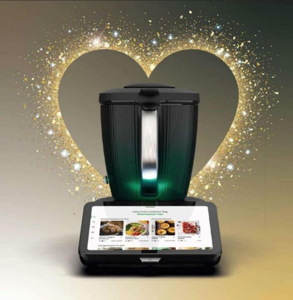 Thermomix® TM7 vor funkelndem Herz aus Goldstaub – leuchtendes Display mit Rezeptübersicht, stilvolle Präsentation für Küchenaktion oder Geschenkidee.