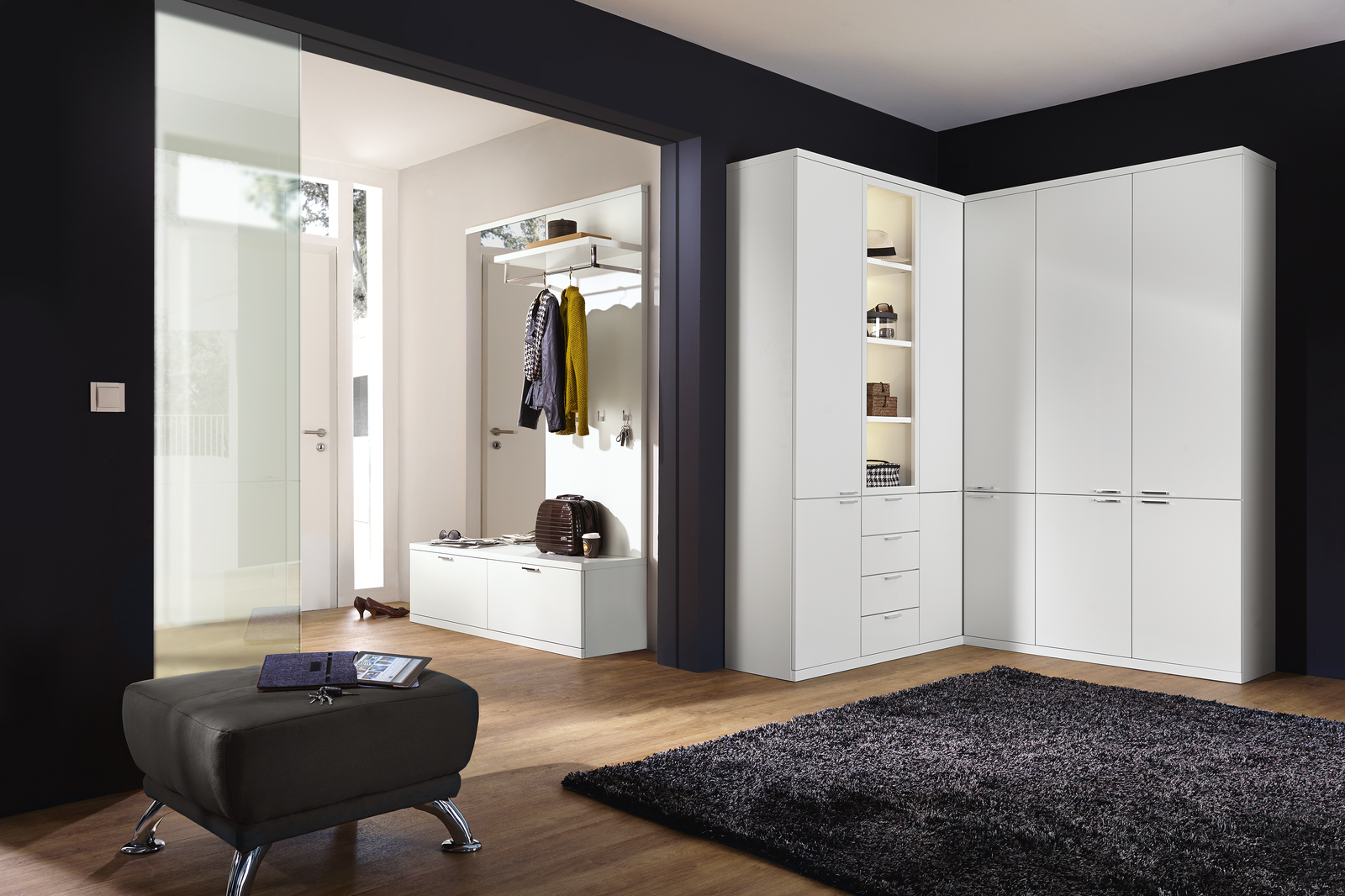 Moderner Eingangsbereich mit weißer Garderobenmöbel-Kombination aus Hochschrank, Sitzbank mit Hakenleiste und offenem Regal. Ein kleiner Hocker mit Tablet und Schlüsseln steht im Vordergrund auf hellem Holzboden. Die dunkle Wand bildet einen eleganten Kontrast, während durch eine große Glastür Tageslicht in den Raum fällt.