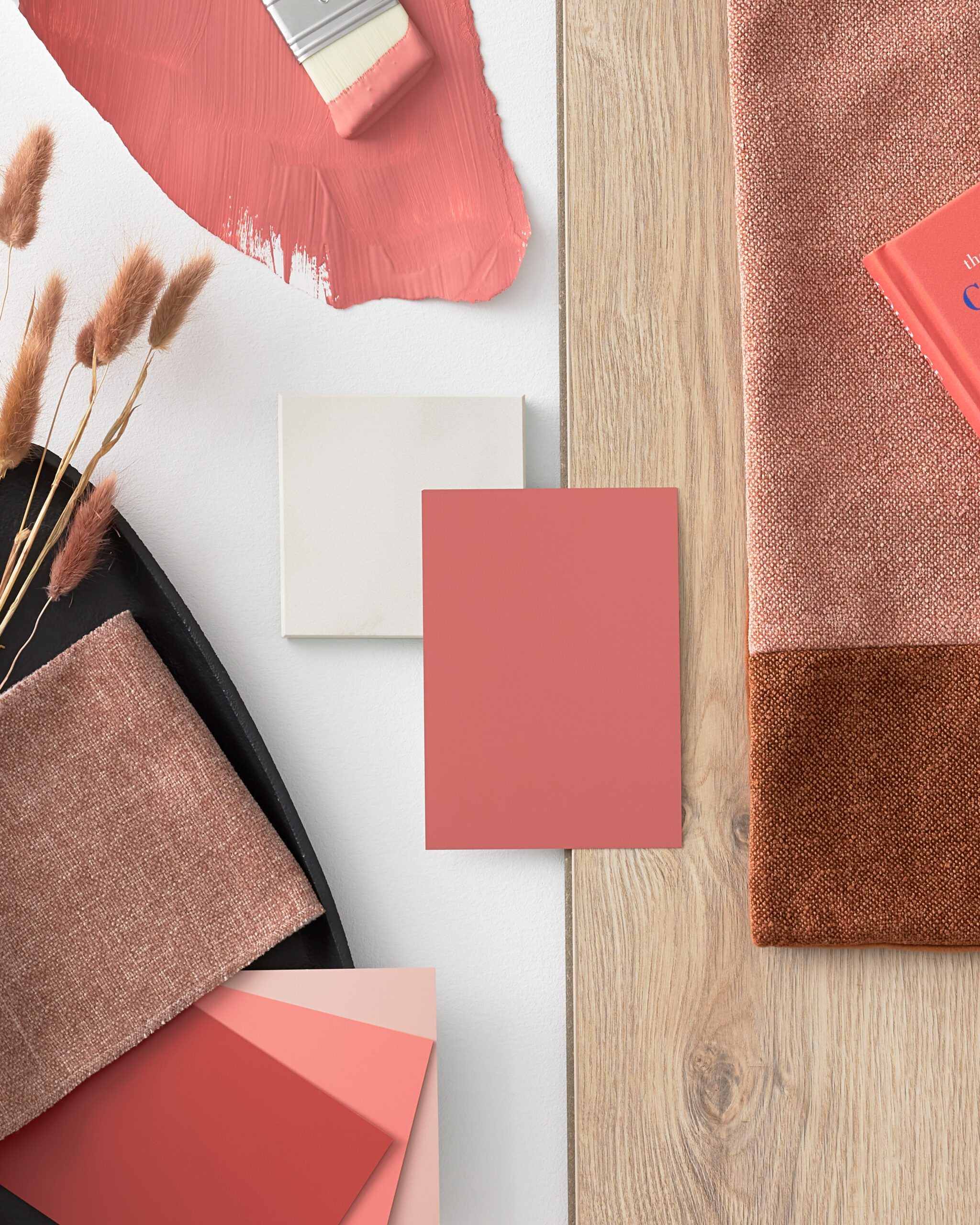 Flachliegende Komposition mit Materialien und Farbmustern in warmen Rosé- und Brauntönen. In der Mitte liegt ein Pinsel mit korallrosa Farbe auf einem bemalten Farbfeld, daneben eine kleine weiße Fliese und mehrere Farbmusterkarten in Rosa und Koralle. Stoffproben in zartem Rosa, Beige und Dunkelbraun ergänzen die Texturvielfalt. Rechts befindet sich ein schwarzes Tablett mit getrockneten Pflanzen und weiteren Textilproben. Eine helle Holzoberfläche bildet den Hintergrund der Szene. Die gesamte Anordnung wirkt wie ein Moodboard für ein Interior- oder Farbgestaltungsprojekt und strahlt eine ruhige, elegante Atmosphäre aus.