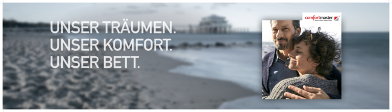 Werbeanzeige mit Strandmotiv – im Hintergrund ist ein Holzsteg (Pier) zu sehen, der ins Meer hinausführt. Links im Bild steht in weißer Schrift auf transparentem Hintergrund der Slogan: „UNSER TRÄUMEN. UNSER KOMFORT. UNSER BETT.“ Rechts ist ein kleineres Bild mit einem sich umarmenden Paar eingeblendet. Im oberen rechten Bereich befindet sich das Logo und der Markenname „comfortmaster“. Die Gesamtgestaltung vermittelt Ruhe, Geborgenheit und Wohlfühlambiente – passend für eine Kampagne rund um komfortables Wohnen oder Schlafen.