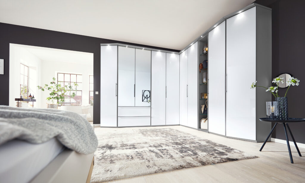 Modernes Schlafzimmer mit weißen Spiegelschränken in L-Form, stilvollem Teppich und Schlafbereich mit grauer Tagesdecke – durchdachtes Stauraumkonzept und elegantes Interior-Design mit natürlichem Licht.