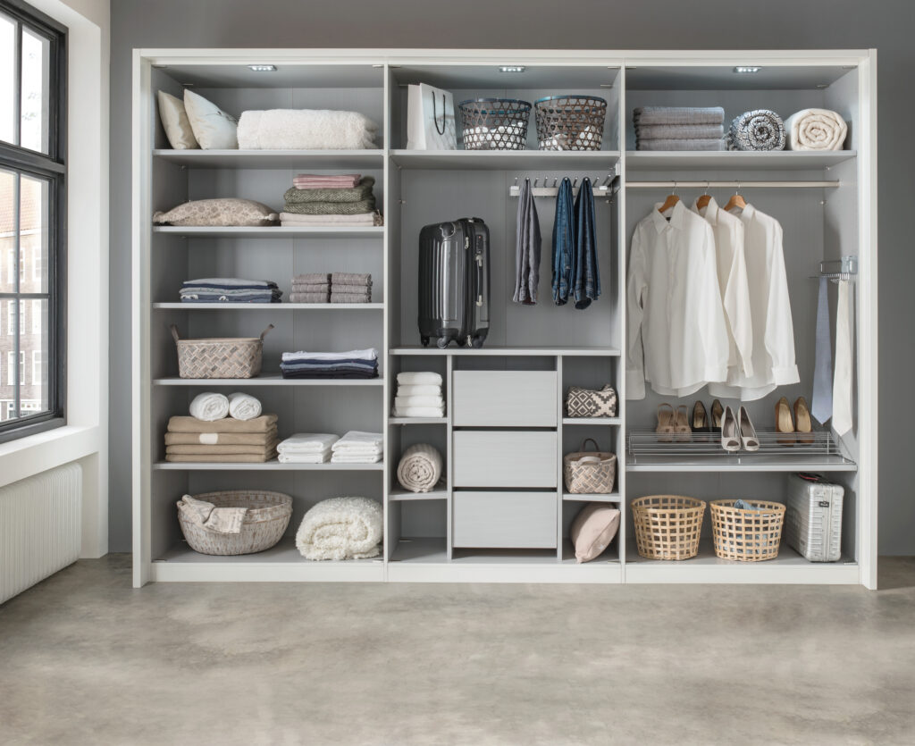Offener Kleiderschrank mit organisiertem Stauraum – gefaltete Kleidung, Handtücher und Boxen auf Regalen, Kleidung ordentlich an Kleiderstangen gehängt, Schuhe auf Drahtregal und ein Koffer am Boden, natürlich beleuchtet durch Fenster – effizientes Ordnungssystem im modernen Wohnraum.