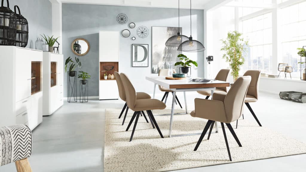 Gemütlich und stilvoll eingerichteter Essbereich mit rechteckigem Tisch, umgeben von vier beigen Polsterstühlen. Über dem Tisch hängen zwei schwarze Pendelleuchten mit Netzschirm-Design. Auf dem Tisch stehen dekorative Objekte wie ein Tablett, eine kleine Pflanze, Geschirr und ein Getränkeglas. Im Hintergrund befinden sich zwei weiße Vitrinenschränke mit Glastüren und eine hohe Kommode mit offenen Regalfächern, die mit weiteren Dekoartikeln bestückt sind. Zwischen den Möbeln stehen zwei schwarze Pflanzenständer mit Grünpflanzen. Die Wand ist mit mehreren dekorativen Elementen versehen – darunter ein großes Poster mit dem Text „Ain’t No Mountain High Enough“, der Schriftzug „Dream“ und geometrische Drahtskulpturen. Rechts fällt durch ein großes Fenster Tageslicht ein. Auf dem Boden liegt ein heller gewebter Teppich.