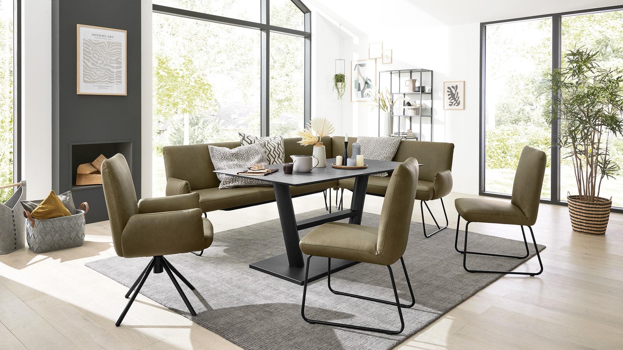 Modernes Esszimmer mit rechteckigem Tisch in dunkler Optik und schwarzem Gestell, umgeben von sechs Stühlen mit olivgrüner Polsterung und schwarzen Metallbeinen. Eine passende Bank mit olivgrünem Bezug und gemusterten Kissen steht an der Längsseite. Der Raum ist hell durch große Fenster mit Blick ins Grüne. Links ein Kamin mit Holzscheiten und darüber ein gerahmtes Bild mit der Aufschrift ‚MODERN ART‘. Daneben ein Korb mit Kissen. Rechts ein Regal mit Dekoration, Büchern und Vasen sowie eine große Zimmerpflanze. Der Boden ist aus hellem Holz, unter dem Tisch liegt ein grauer Teppich.