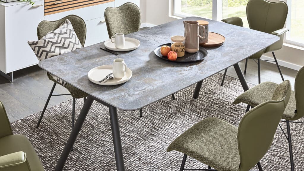Modernes Esszimmer mit rechteckigem Tisch in grauer Steinoptik, gedeckt mit zwei Tellern, Tassen und Besteck. Auf dem Tisch steht ein schwarzes Tablett mit einem Krug, einem Holzbehälter und drei Äpfeln. Sechs schlanke Stühle mit grünlicher Polsterung umgeben den Tisch, einer davon mit gemustertem Kissen. Der Raum ist hell durch ein großes Fenster, mit einem gewebten Teppich am Boden und einem weißen Sideboard mit Holzakzenten im Hintergrund.