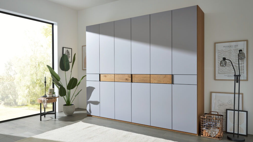 Zeitgemäß gestalteter Wohnraum mit großem grauen Kleiderschrank in Holzoptik, dekorativem Beistelltisch mit Kerzen und Pflanzen vor einem Fenster, ergänzt durch moderne Wandkunst und Stehlampe – minimalistisches Interior-Design mit natürlichen Akzenten.