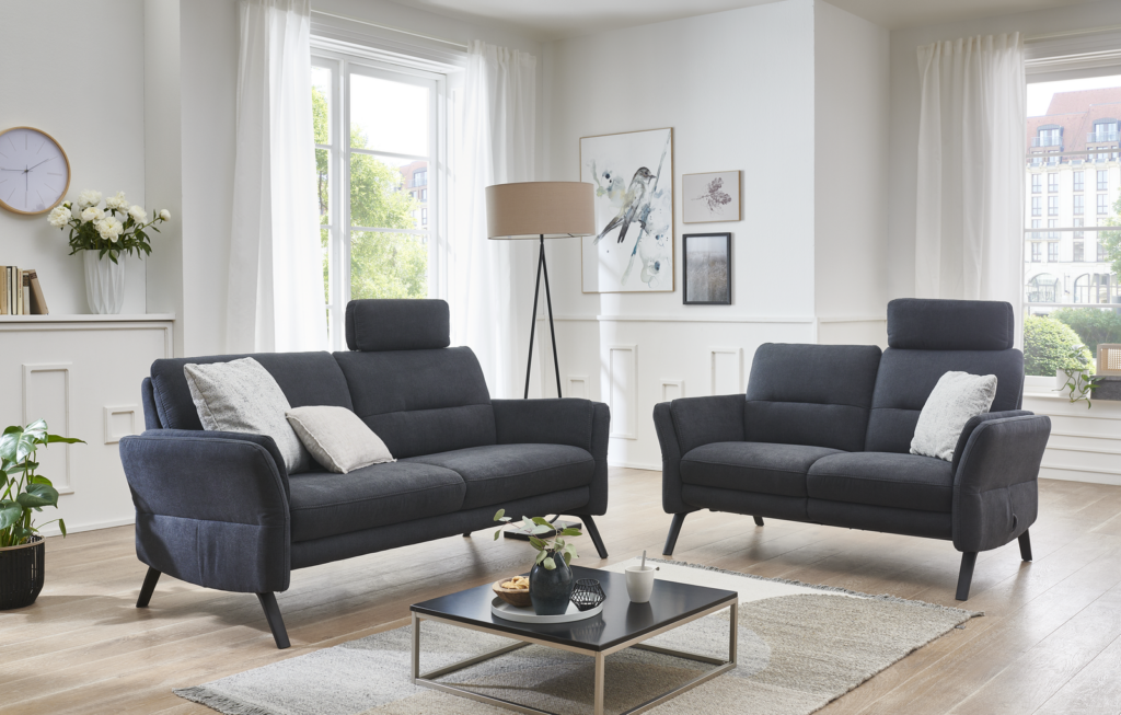 Ein geschmackvoll eingerichtetes Wohnzimmer mit zwei dunkelgrauen Sofas, die sich gegenüberstehen. In der Mitte steht ein rechteckiger Couchtisch mit einer grünen Pflanze und einem Tablett. Der Raum wird von großen Fenstern mit weißen Vorhängen erhellt, wodurch viel Tageslicht einfällt. An der Wand hängen gerahmte Kunstwerke, eine Uhr sowie ein Regal mit einer Vase voller Blumen. Eine schlichte Stehlampe steht in der Ecke. Das Design wirkt modern und gemütlich mit klaren Linien und einer neutralen Farbpalette.