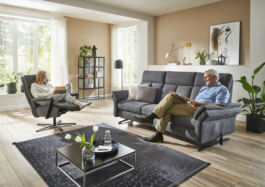 Ein stilvoll eingerichtetes Wohnzimmer mit zwei Personen, die jeweils ein Buch lesen. Eine Person sitzt entspannt in einem schwarzen Drehsessel, die andere auf einem grauen Sofa. Der Raum ist lichtdurchflutet durch große Fenster und wirkt modern und einladend. Schwarze Möbel und Regale mit Zimmerpflanzen, Büchern und Dekoration verleihen dem Raum einen gemütlichen Charme. Auf dem schwarzen Couchtisch stehen eine Vase und ein Glas Wasser. An der Wand hängt ein Kunstwerk, und auf einem weißen Regal steht eine Lampe. Der Holzboden und der dunkle Teppich runden die ruhige Atmosphäre ab.