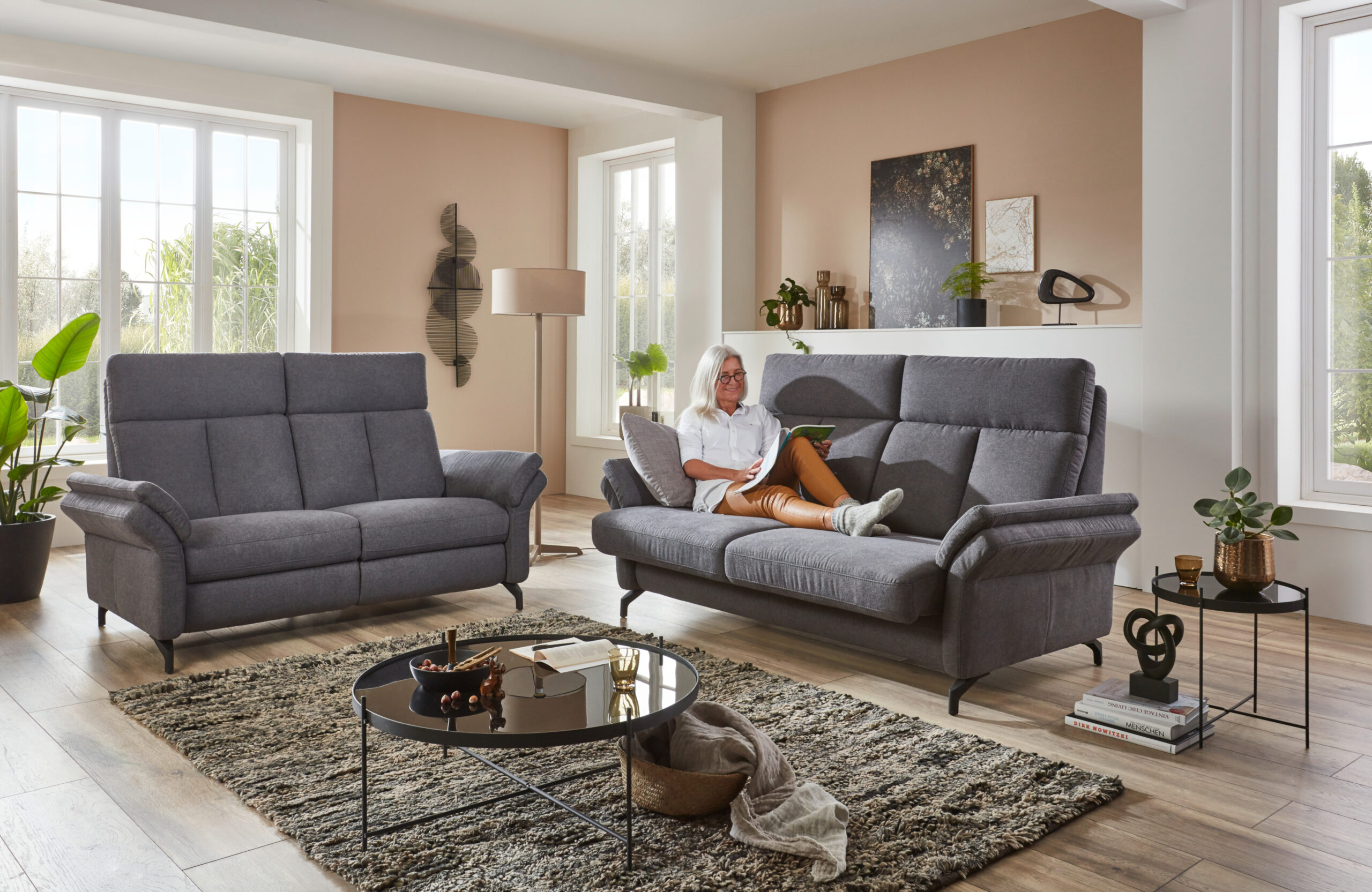 2 große 2-sitzige Hochlehner Sofas in graum Stoff. Eine ältere Frau liegt entspannt auf dem Sofa.