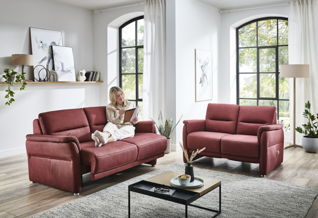 Ein helles, modernes Wohnzimmer mit zwei roten Sofas, die einander gegenüberstehen. Eine Person sitzt entspannt auf dem linken Sofa und hält ein Buch oder Tablet. Große Fenster lassen Tageslicht herein und erhellen den Raum. An der Rückwand hängt ein dekoratives Regal mit Pflanzen, Bildern und Büchern. Auf dem kleinen Couchtisch zwischen den Sofas liegen eine Tasse und ein weiteres Buch. Die Atmosphäre wirkt ruhig und einladend.
