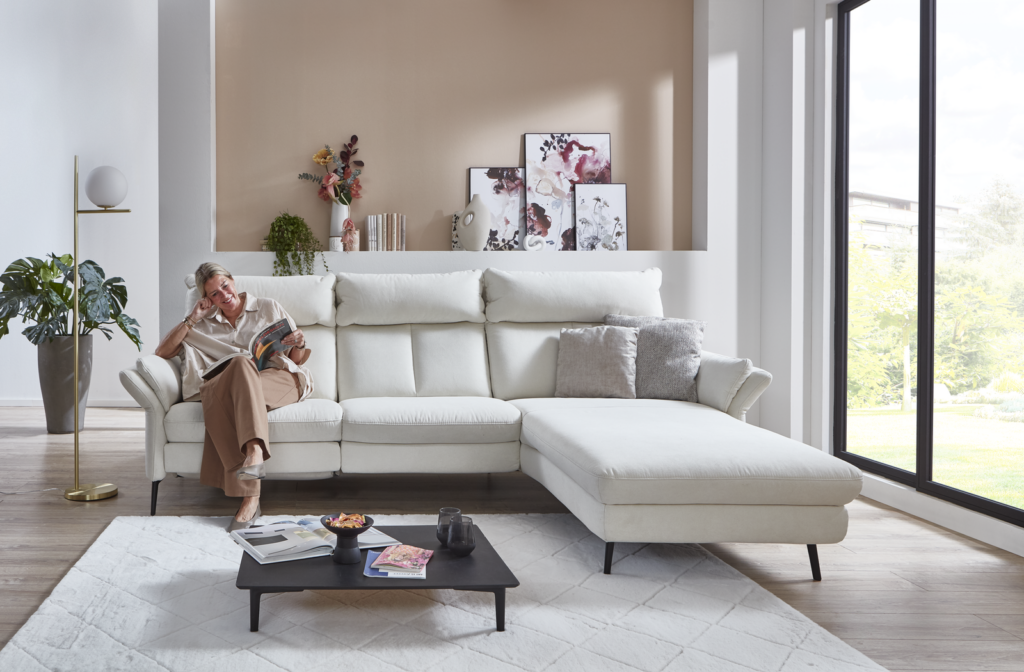 Eine Person sitzt entspannt auf der linken Seite eines großen weißen Ecksofas in einem modern eingerichteten Wohnzimmer und liest ein Buch. Vor dem Sofa steht ein Couchtisch mit Snacks, einer Fernbedienung und Zeitschriften. Durch ein großes Fenster rechts fällt viel Tageslicht in den Raum. Die Wand hinter dem Sofa ist mit Kunstwerken und Pflanzen dekoriert und verleiht dem Raum eine gemütliche Atmosphäre.
