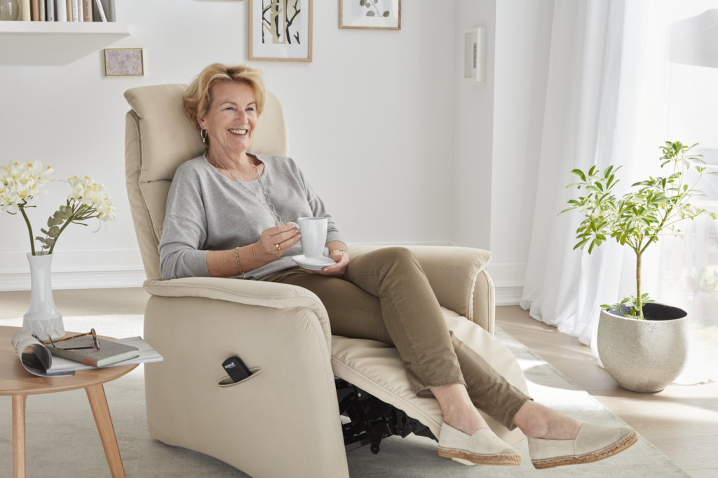 Eine Person sitzt entspannt in einem beigen Relaxsessel in Liegeposition und hält eine weiße Tasse in der Hand. Neben dem Sessel steht ein kleiner Holztisch mit einer Vase, in der weiße Blumen arrangiert sind, sowie ein aufgeklapptes Buch. Im Hintergrund befinden sich zwei gerahmte Bilder an der Wand und ein großer grüner Blumentopf. Der Raum wirkt freundlich und gemütlich mit viel Tageslicht.