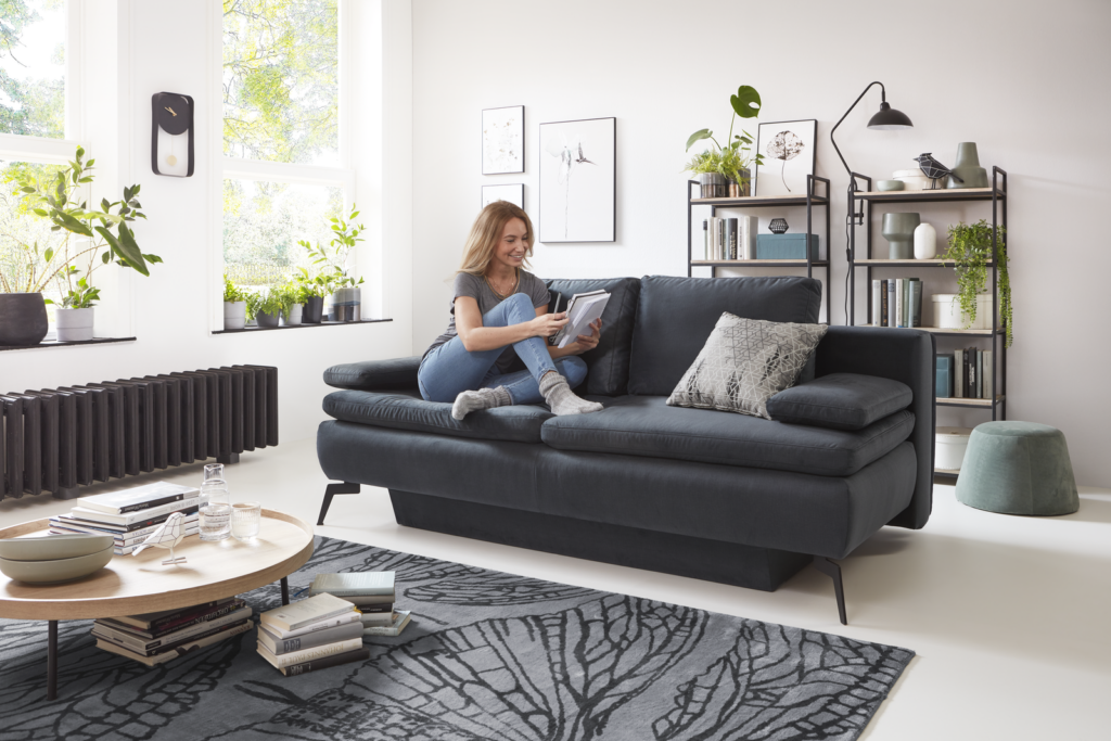 Modern gestaltetes Wohnzimmer mit dunkelgrauem Schlafsofa, auf dem eine Person sitzt und ein Buch liest. Das Sofa ist mit einem gemusterten Kissen dekoriert, davor steht ein runder Holz-Couchtisch mit Schale, Glas und Büchern. Der Boden ist mit einem grauen Teppich bedeckt. Im Hintergrund befinden sich große Fenster mit mehreren Zimmerpflanzen auf der Fensterbank sowie ein schwarzer Heizkörper darunter. Zwei Wandregale mit Büchern und Deko, Bilderrahmen und eine Uhr schmücken die Wand – ein gemütlicher Rückzugsort mit stilvoller Einrichtung