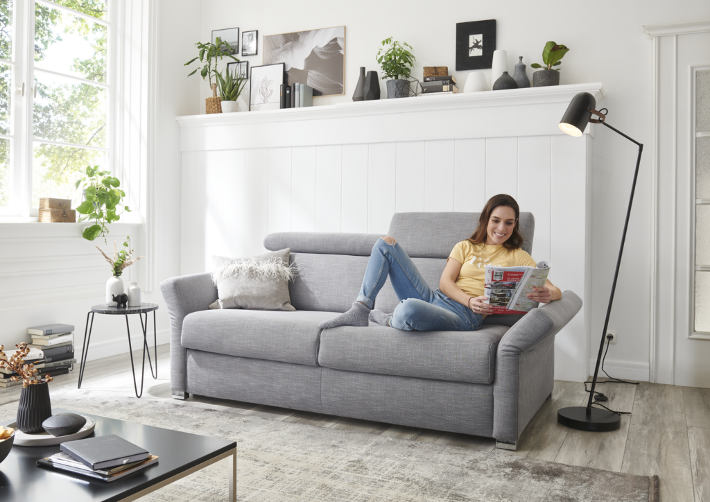 Modern eingerichtetes Wohnzimmer mit grauem Schlafsofa, auf dem eine Person sitzt und eine Zeitschrift liest. Das Sofa steht vor einer weißen Wand mit Regal, das Bücher, Bilderrahmen, Pflanzen und Deko-Objekte beherbergt. Links fällt Tageslicht durch ein großes Fenster mit weißen Vorhängen, auf dessen Fensterbank eine Zimmerpflanze steht. Ein runder Beistelltisch mit weißer Vase und Pflanze sowie eine schwarze Stehlampe ergänzen die Einrichtung. Im Vordergrund befindet sich ein dunkler Couchtisch mit Büchern und Dekoration auf einem hellen Teppich – eine gemütliche Kombination aus Stil und Funktion.