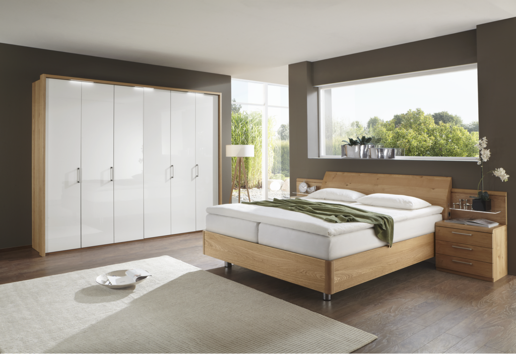 Modernes Schlafzimmer mit großem Holzbett, weißer Bettwäsche und grüner Tagesdecke – rechts daneben Holz-Nachttisch mit Uhr und Dekoration. Hinter dem Bett großes Fenster mit Blick ins Grüne, Fensterbank mit zwei Pflanztöpfen. Links hoher weißer Kleiderschrank mit silbernen Griffen, daneben Stehleuchte. Heller Teppich auf Holzfußboden – ruhige, lichtdurchflutete Schlafzimmerszene mit natürlichem Flair.