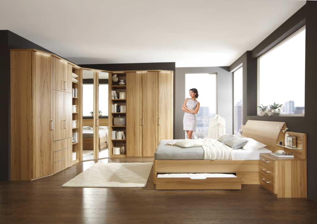 Modernes Schlafzimmer mit großem Holzbett und Schublade unter der Liegefläche, Holz-Nachttisch mit drei Schubladen und kleiner Ablagefläche – dekoriert mit Uhr und Buch. Im Hintergrund raumhohe Fenster mit Blick auf eine Stadtlandschaft. Links eingebauter Holz-Kleiderschrank mit offenen Regalfächern, Schubladen und Spiegel. Bücher und Deko-Objekte setzen wohnliche Akzente – durchdachte Einrichtungslösung mit viel Stauraum und hellem Holzfußboden.