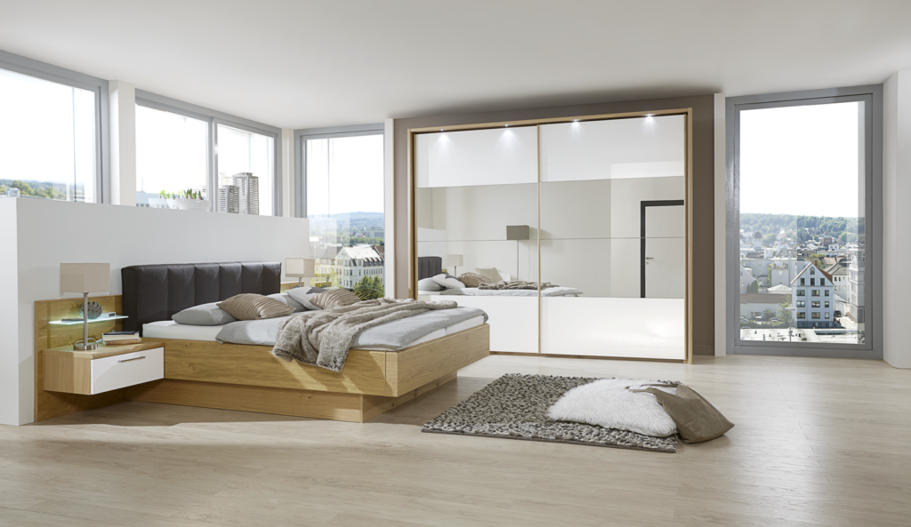 Modernes Schlafzimmer mit großem Holz-Doppelbett und gepolstertem Kopfteil – dekoriert mit weißen und grauen Kissen sowie beigefarbener Decke. Links und rechts jeweils Nachttisch mit Lampe. Im Hintergrund große Fenster mit Blick auf Stadtgebäude, lassen viel Tageslicht hinein. Gegenüber vom Bett großer Kleiderschrank mit Spiegelfronten, die den Raum reflektieren. Auf dem hellen Holzfußboden liegt ein strukturierter Teppich mit Kissen und weißer Kuscheldecke – stilvolles, urbanes Ambiente mit klarer Linienführung