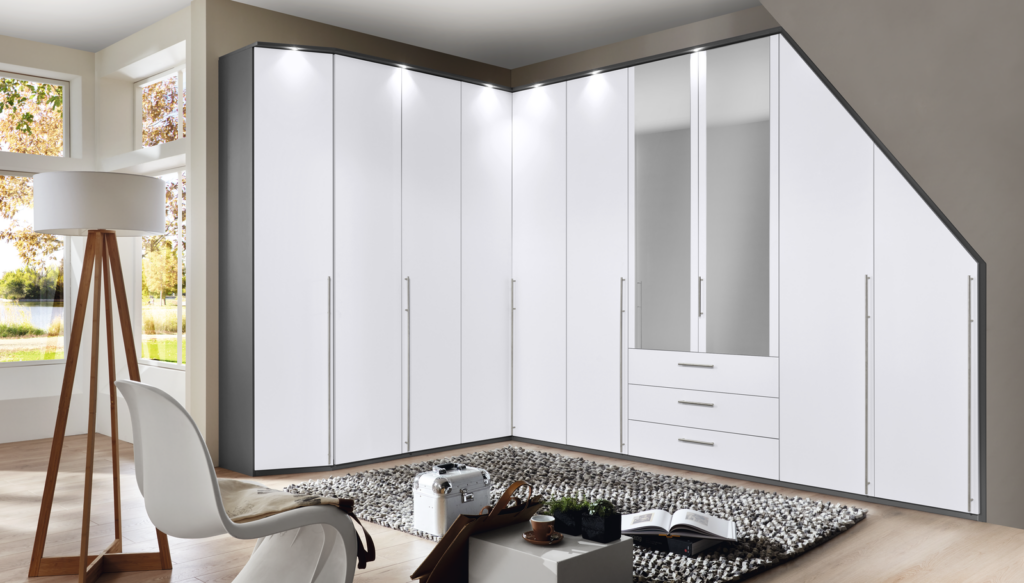Modernes, lichtdurchflutetes Schlafzimmer mit großem, weißem Eck-Kleiderschrank in L-Form – ausgestattet mit Spiegeltüren, Schubladen und langen Griffleisten. Vor dem Schrank weiße Design-Chaiselongue mit beiger Decke, daneben Holz-Stativlampe mit weißem Schirm. Runder Beistelltisch mit Tasse, Buch und Deko-Elementen auf grauem Teppichboden. Großes Fenster mit Blick auf grüne Bäume und Gewässer – klares, ruhiges Raumkonzept mit eleganter Stauraumlösung