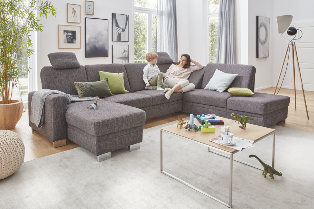 Familienfreundliches Wohnzimmer mit großem, grauem Ecksofa, auf dem ein Erwachsener und ein Kind sitzen. Das Sofa ist mit Kissen in Gelb und Grün dekoriert. Vor dem Sofa steht ein Couchtisch mit Spielzeug, Büchern und einem Dinosaurier-Modell, was auf eine entspannte Spielzeit hinweist. Der Raum ist durch große Fenster lichtdurchflutet und mit Vorhängen versehen. Eine große Zimmerpflanze, eine dreibeinige Stehlampe und gerahmte Bilder an der Wand sorgen für eine gemütliche, lebendige Atmosphäre.