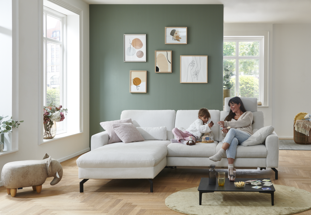 Gemütliches Wohnzimmer mit weißem Ecksofa, auf dem ein Erwachsener und ein Kind sitzen. Der Erwachsene hält eine Tasse, während das Kind aus einer Schüssel isst. Hinter dem Sofa befindet sich eine grüne Wand mit fünf gerahmten Bildern, die abstrakte und minimalistische Motive zeigen. Links sorgt ein großes Fenster für viel Tageslicht. Vor dem Sofa steht ein schwarzer Couchtisch mit Getränken und Snacks. Ein kleiner hölzerner Hocker in Elefantenform rundet die verspielte Atmosphäre ab. Der Raum wirkt hell, freundlich und familiär.