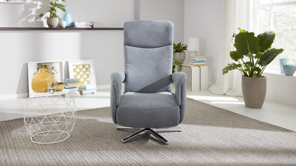 Modernes Wohnzimmer mit einem grauen Relaxsessel auf Drehfuß, der zentral auf einem grauen Teppich steht. Links neben dem Sessel befindet sich ein weißer Beistelltisch mit Glasplatte, auf dem ein gelbe Vase, eine kleine weiße Schale und ein Dekogegenstand arrangiert sind. Im Hintergrund hängt ein weißes Wandregal mit Büchern, einer kleinen Pflanze und einem gerahmten Bild. Rechts steht eine große Zimmerpflanze mit breiten grünen Blättern in einem weißen Topf. Durch weiße Vorhänge fällt Tageslicht in den Raum, das die minimalistische und luftige Gestaltung unterstreicht.