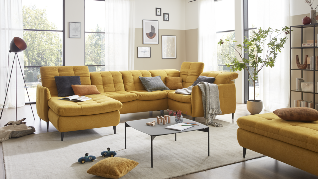 Modernes Wohnzimmer mit einem großen, modularen Ecksofa in leuchtendem Gelb, ergänzt durch passende gelbe Hocker. Auf dem zentral stehenden Couchtisch befindet sich ein dekoratives Schachspiel sowie weitere Accessoires. Graue und blaue Kissen sorgen für Farbakzente und Komfort. Der Raum wird durch große Fenster mit transparenten Vorhängen hell erleuchtet. Eine Stehlampe mit braunem Schirm steht links neben dem Sofa, rechts schmückt eine Zimmerpflanze den Bereich am Fenster. Die Wände sind mit gerahmten Bildern und Fotografien dekoriert, was dem Raum eine persönliche und wohnliche Note verleiht.