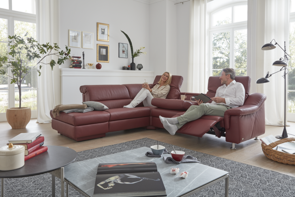 Zwei Personen sitzen entspannt auf einem roten Leder-Ecksofa in einem modern eingerichteten Wohnzimmer. Der Raum ist lichtdurchflutet, mit großen Fenstern und weißen Vorhängen. Links steht eine große Zimmerpflanze, rechts eine stilvolle Stehleuchte mit mehreren verstellbaren Leuchtelementen. Die Rückwand ziert ein weißes Wandregal mit dekorativen Objekten wie Büchern, Bildern und Vasen. Vor dem Sofa steht ein Couchtisch mit einem offenen Buch, zwei Schalen und kleinen Dekoartikeln. Der Boden ist von einem grafisch gemusterten Teppich bedeckt – ein gemütliches Ambiente mit klarer Komfortbotschaft.