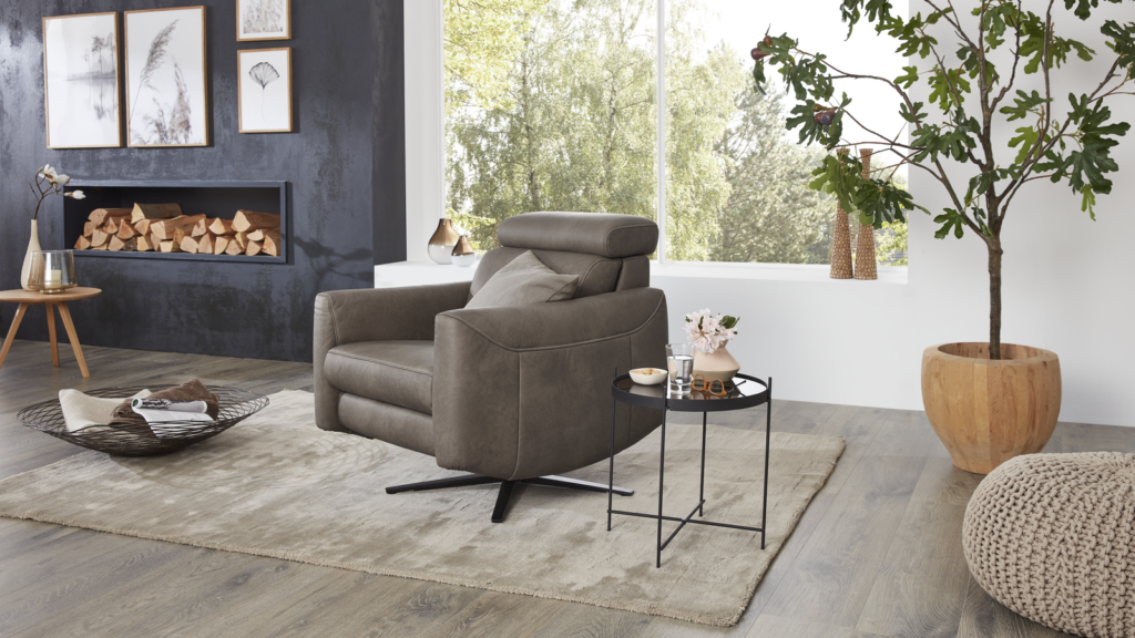 Grauer Comfortmaster-Relaxsessel mit Kissen auf hellem Teppich, daneben schwarzer runder Beistelltisch mit Wasserglas, Schale und Vase mit Blumen. Im Hintergrund dunkle Wand mit drei gerahmten Bildern und Regal mit gestapeltem Brennholz. Links Holztisch mit Deko-Vase, rechts große Zimmerpflanze und gestrickter Pouf – stilvolles Wohnzimmer mit natürlichem Licht und Blick ins Grüne