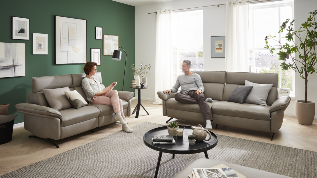 Modernes Wohnzimmer mit zwei Personen auf gegenüberliegenden Ledersofas, dazwischen ein runder Couchtisch mit Teekanne, Tassen und Buch. Der Raum ist stilvoll dekoriert mit grüner Akzentwand, mehreren gerahmten Wandbildern, weißer Stehlampe und großer Zimmerpflanze. Tageslicht fällt durch große Fenster mit weißen Vorhängen auf hellen Holzfußboden und grauen Teppich – eine Szene mit entspannter Stimmung und stilvoller Wohnästhetik.