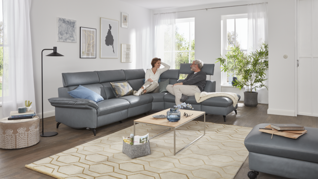 Großzügiges Wohnzimmer mit grauem Ecksofa, auf dem zwei Personen sitzen – eine mit blauem Zierkissen im Rücken, die andere mit einem gemusterten Kissen. Vor dem Sofa liegt ein Teppich mit hexagonalem Muster, darauf ein Holz-Couchtisch mit schwarzen Metallbeinen, dekoriert mit einer Vase mit Blumen und Glasobjekten. Links daneben steht ein runder Hocker mit Büchern und Pflanze sowie eine schwarze Stehlampe. Der Hintergrund zeigt eine Wand mit mehreren gerahmten Kunstmotiven und großen Fenstern mit weißen Vorhängen, die Tageslicht hereinlassen. Rechts im Bild eine üppige Zimmerpflanze und ein grauer Hocker mit Notizbuch – ein wohnliches Ambiente zwischen Gemütlichkeit und moderner Designästhetik