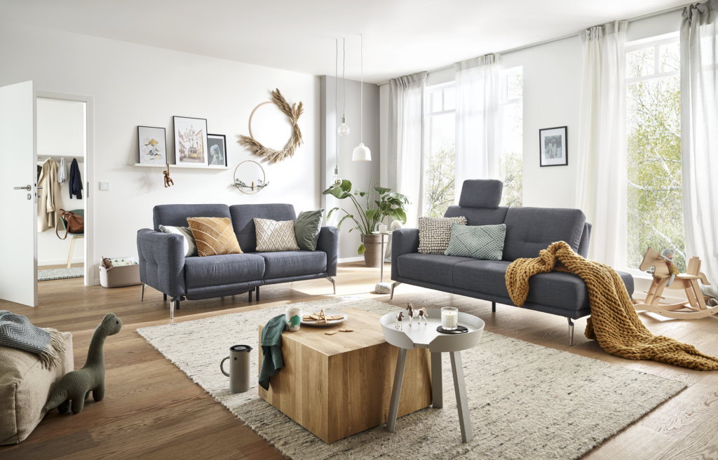 Gemütlich eingerichtetes Wohnzimmer mit zwei grauen Sofas, dekoriert mit bunten Kissen. In der Mitte steht ein Holztisch mit einem Schachspiel, einer Kerze und einer Tasse. Der Raum wirkt hell und freundlich dank großer Fenster mit transparenten Vorhängen. Auf dem Boden liegt ein heller Teppich, umgeben von Spielzeug wie einem Dinosaurier und einer Gießkanne. Pflanzen, gerahmte Bilder und ein Kranz an der Wand verleihen dem Raum eine warme, einladende Atmosphäre.