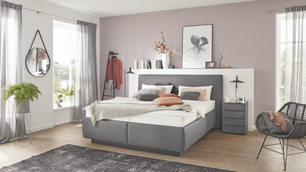 Modernes Schlafzimmer in beruhigenden Grau- und Rosatönen. Das Bett verfügt über einen gepolsterten Rahmen und ein graues Kopfteil, dekoriert mit verschiedenen Kissen in Weiß, Grau und Rosa. Neben dem Bett steht ein grauer Nachttisch mit Lampe, Tasse und gerahmtem Bild. Über dem Bett befindet sich ein Wandregal mit Büchern, Dekoästen und Kunstwerk. Links im Raum: großes Fenster mit grauen Vorhängen, runde Wandspiegel, grüne Pflanze auf einem Hocker sowie eine schwarze Leiter mit hängendem Kleidungsstück. Rechts: schwarzer Korbstuhl mit rosa Stoff. Holzfußboden und dunkler Teppich runden die gemütliche, stilvolle Atmosphäre ab.