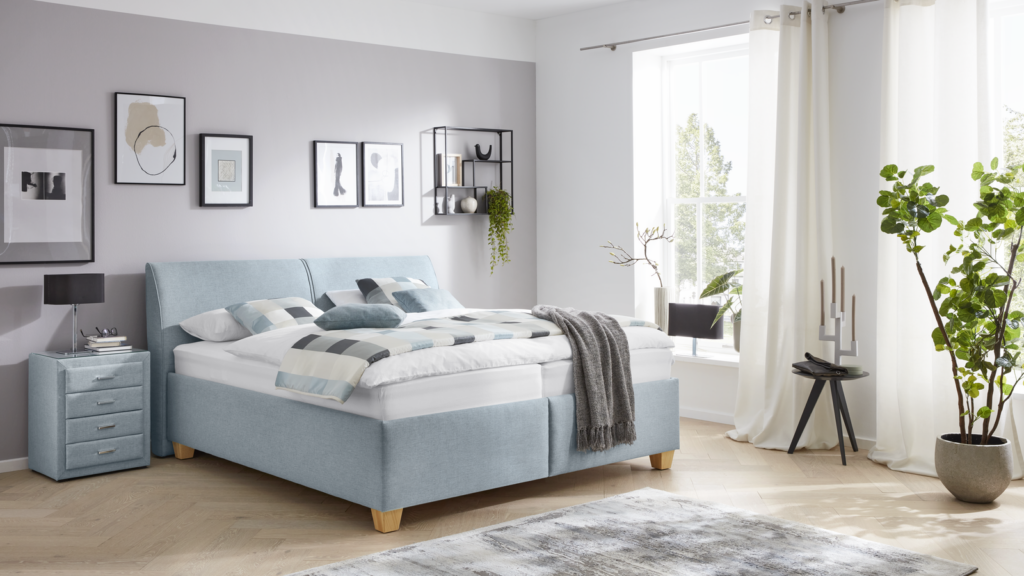 Modern eingerichtetes Schlafzimmer mit einem hellblauen, gepolsterten Bett auf Holzfüßen. Das Bett ist mit einem karierten Bettbezug und passenden Kissen ausgestattet, darüber liegt eine graue Decke. Links steht ein kleiner Nachttisch mit vier Schubladen, einem schwarzen Tischlampe und einigen Büchern. Über dem Bett hängen vier gerahmte Bilder sowie ein schwarzes Wandregal mit Dekoartikeln und einer hängenden Pflanze. Rechts sorgt ein großes Fenster mit weißen Vorhängen für Tageslicht. Vor dem Fenster stehen ein runder Beistelltisch mit einem Kerzenständer sowie eine grüne Topfpflanze auf dem Boden. Der Raum wirkt klar, minimalistisch und einladend.