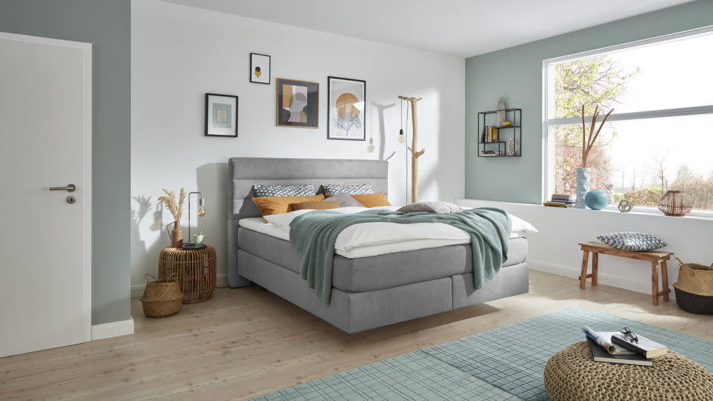 Gemütlich eingerichtetes Schlafzimmer mit einem großen, grau gepolsterten Bett. Das Bett ist mit weißen Textilien, grauen und senfgelben Kissen sowie einer grünen Decke dekoriert. Über dem Bett hängen vier gerahmte Bilder. Links steht ein geflochtener Beistelltisch mit einer Vase voller Trockenblumen, daneben ein hängendes Leuchtmittel und ein Korb am Boden. Rechts befindet sich ein Holz-Kleiderständer mit Leuchte. Vor dem Fenster mit Blick ins Grüne steht eine Holzbank mit Kissen, Vase und Büchern. Auf dem Boden liegen ein gemusterter Teppich und ein gewebter Pouf mit Brille und Buch. Der Raum wirkt modern, hell und sehr einladend.