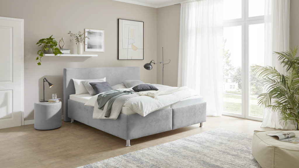 Modernes Schlafzimmer mit hellgrauem Polsterbett, das mit weißer und grauer Bettwäsche sowie mehreren Kissen dekoriert ist. Links neben dem Bett steht ein runder Beistelltisch mit schwarzer Lampe, Trinkglas und Büchern. Darüber befindet sich ein weißes Wandregal mit Zimmerpflanze, Dekoobjekt und weiteren Büchern. Über dem Bett hängen zwei gerahmte Kunstwerke – ein abstraktes Bild und ein Schwarz-Weiß-Foto. Rechts im Raum steht eine schwarze Stehlampe. Große Fenster mit weißen Vorhängen lassen Tageslicht hinein, daneben eine hohe grüne Pflanze. Der Holzfußboden und ein heller Teppich sorgen für eine warme, gemütliche Atmosphäre.