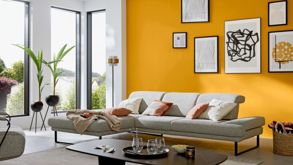 Graues Ecksofa von Interliving 4460 vor gelber Wand. Schön Dekoriert mit ein paar Bildern.