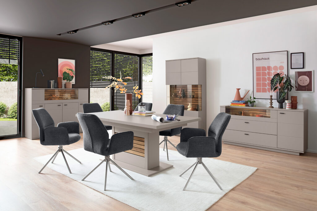 Modernes Esszimmer mit großem rechteckigem Tisch und fünf grauen, gepolsterten Drehstühlen mit Metallbeinen. Der Raum ist mit hellen Holzböden und einem weißen Teppich unter dem Tisch ausgestattet. Im Hintergrund stehen drei Aufbewahrungsmöbel mit Kombinationen aus geschlossenen und gläsernen Fronten sowie Holzdetails. Wände sind mit gerahmten Kunstwerken dekoriert, darunter ein Bauhaus-Poster und ein pflanzenbezogenes Druckmotiv. Große Fenster mit schwarzen Jalousien lassen Tageslicht hinein und bieten Blick auf grünes Außengelände. Dekorationen wie Vasen, Bücher und Pflanzen verleihen dem Raum Persönlichkeit.