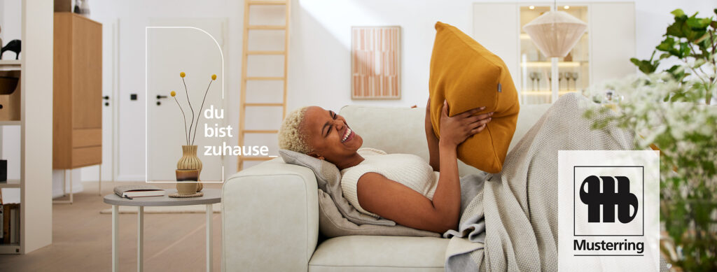 Wohnzimmer-Szene mit Person auf grauem Sofa, umgeben von moderner Einrichtung und dekorativen Elementen – gemütlicher Rückzugsort mit Stil, Pflanzen und warmem Licht, ergänzt durch den Schriftzug ‚du bist zuhause‘.