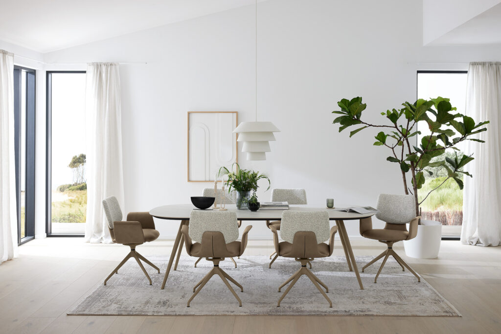 Helles, modern eingerichtetes Esszimmer mit ovalem Tisch und sechs beigen Stühlen, dekoriert mit Kerzen, Pflanzen und Magazinen. Über dem Tisch hängt eine stilvolle, weiße Pendelleuchte. Der Raum wird durch große Fenster mit weißen Vorhängen von Tageslicht durchflutet. Ein großes Pflanzenarrangement und abstrakte Kunst an der Wand vervollständigen das ruhige, zeitlose Design.
